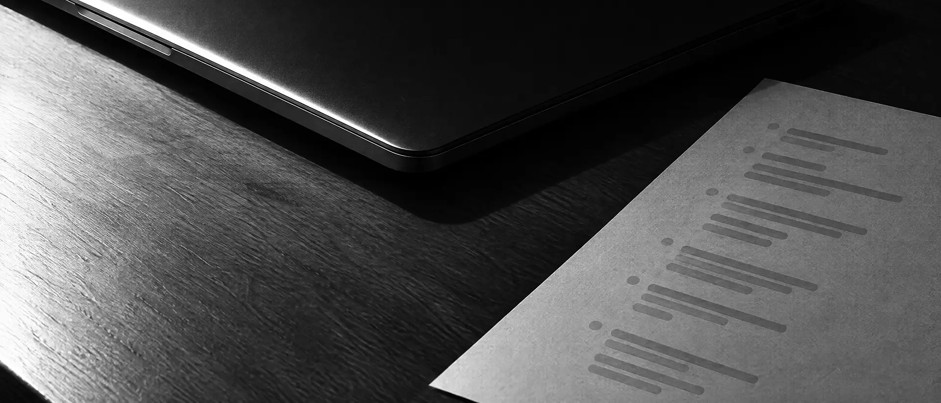 Geschlossener Laptop mit dunklem Deckel auf einem Holztisch, daneben ein bedrucktes Blatt mit Zeilen