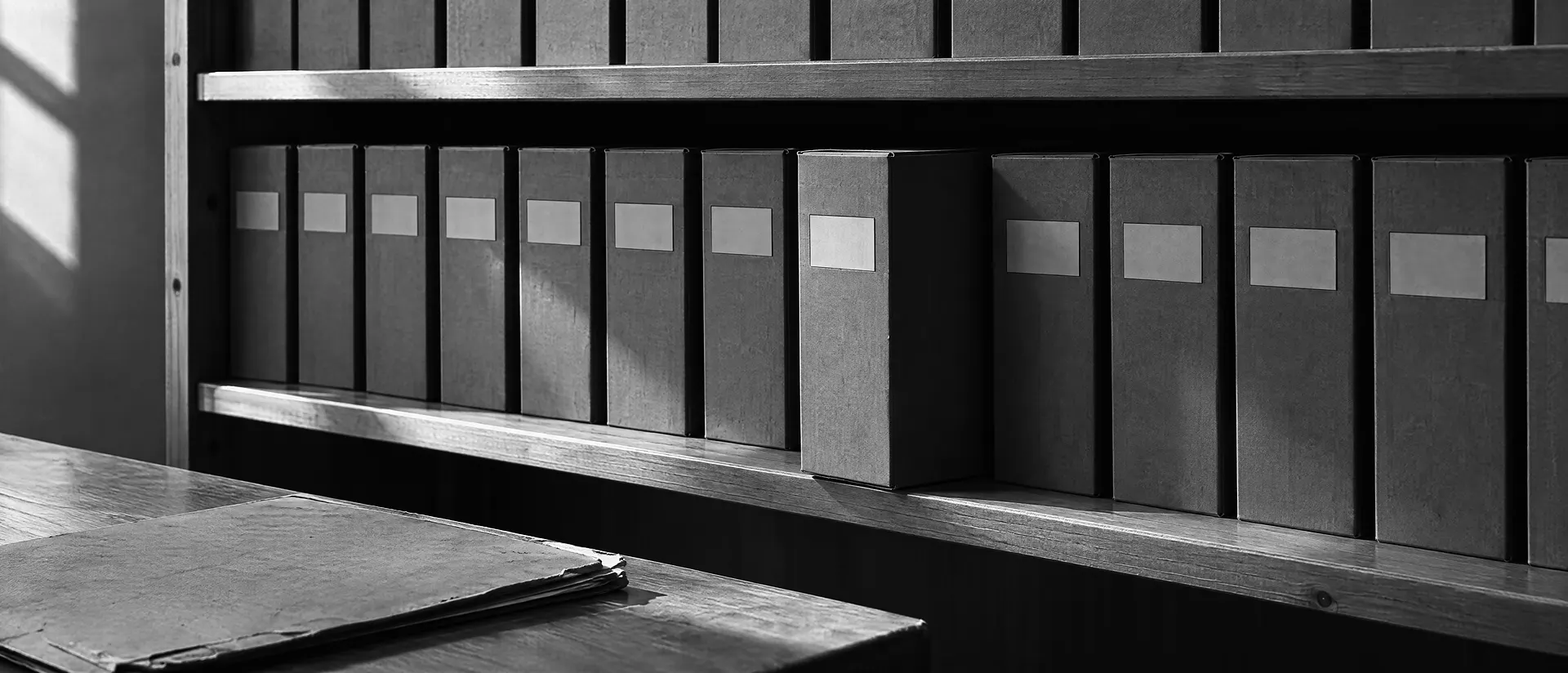 Archivregal mit einer Reihe von Kartons, deren Beschriftungen leer und unleserlich sind, davor ein abgegriffener Aktenband