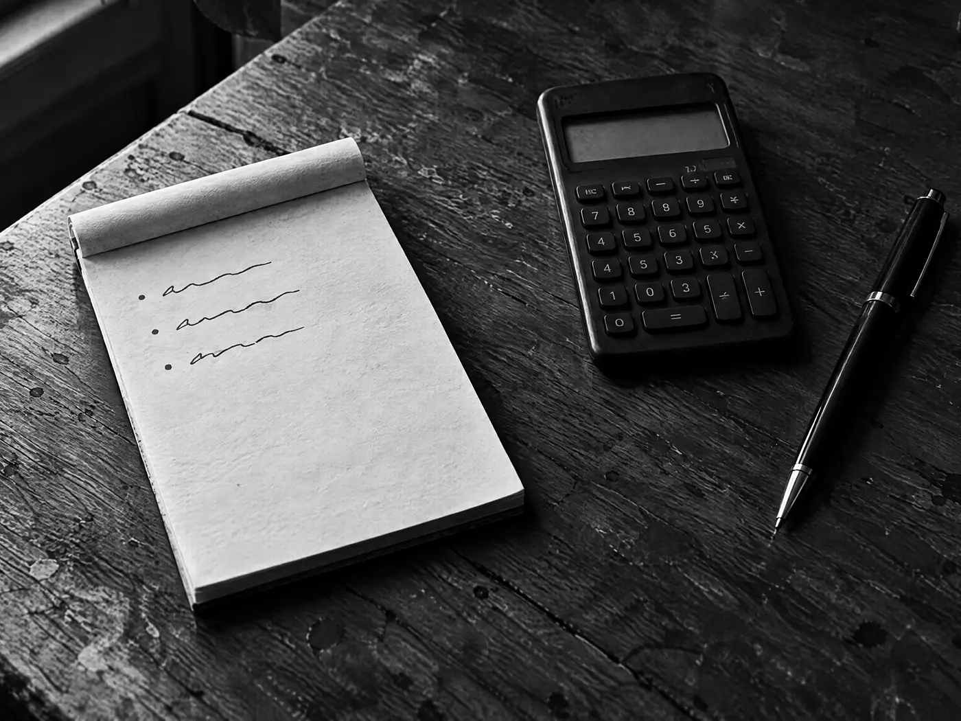 Notizblock mit unleserlichen handschriftlichen Zeilen, daneben ein Taschenrechner mit dunklem Display und ein Stift auf einem Holztisch