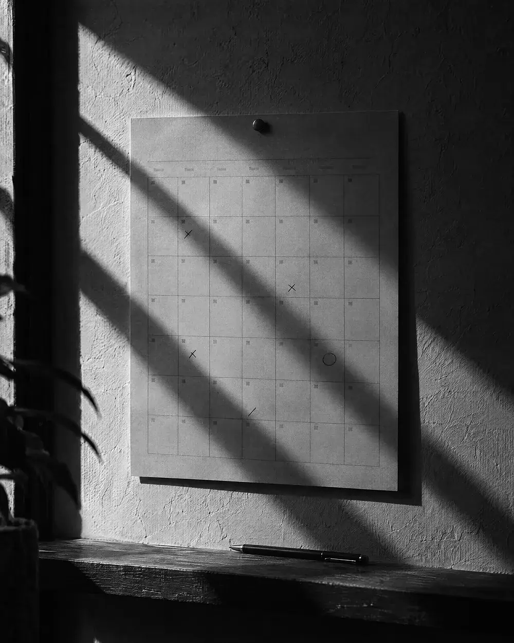 Wandkalender mit einzelnen markierten Feldern an einer verputzten Wand, darunter ein Stift im harten Streiflicht