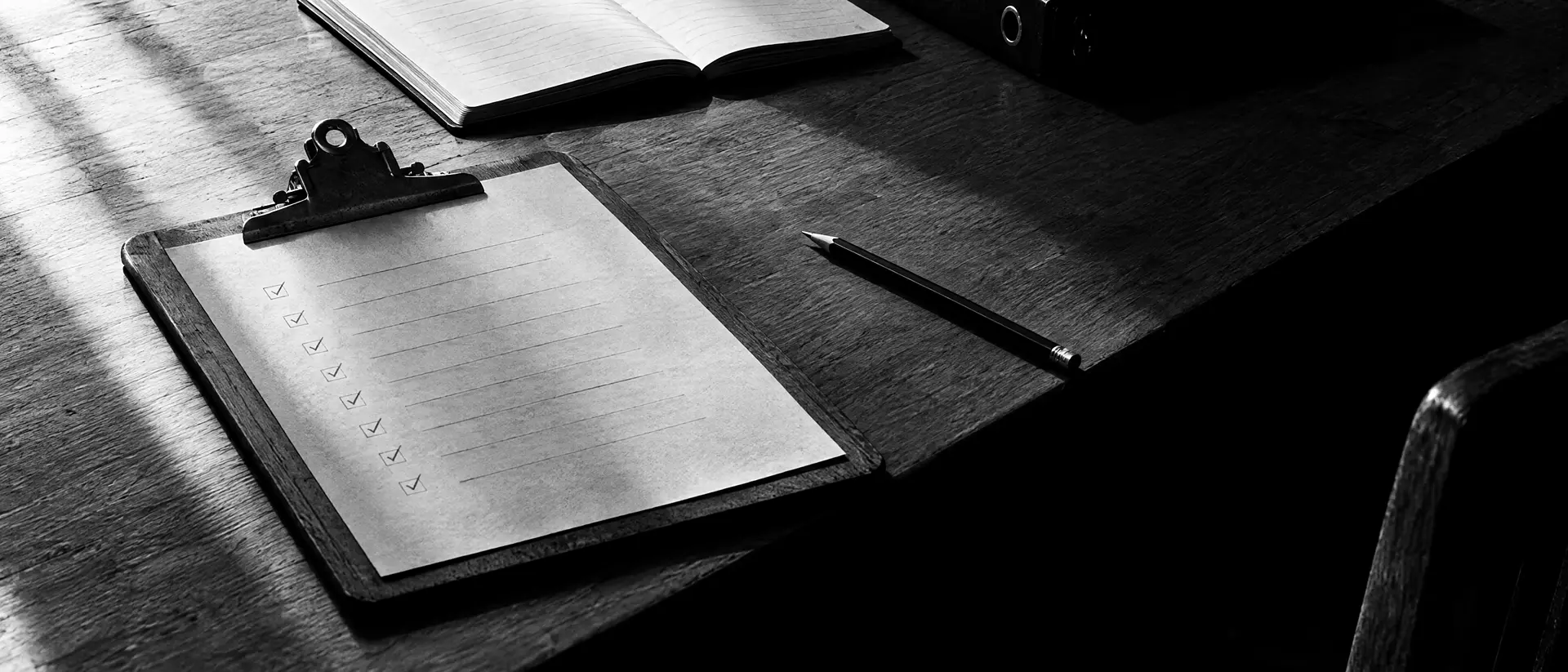 Klemmbrett mit einer Liste kleiner Ankreuzfelder und einem Bleistift, daneben ein aufgeschlagenes Notizbuch und ein geschlossener Ordner auf einem Holztisch
