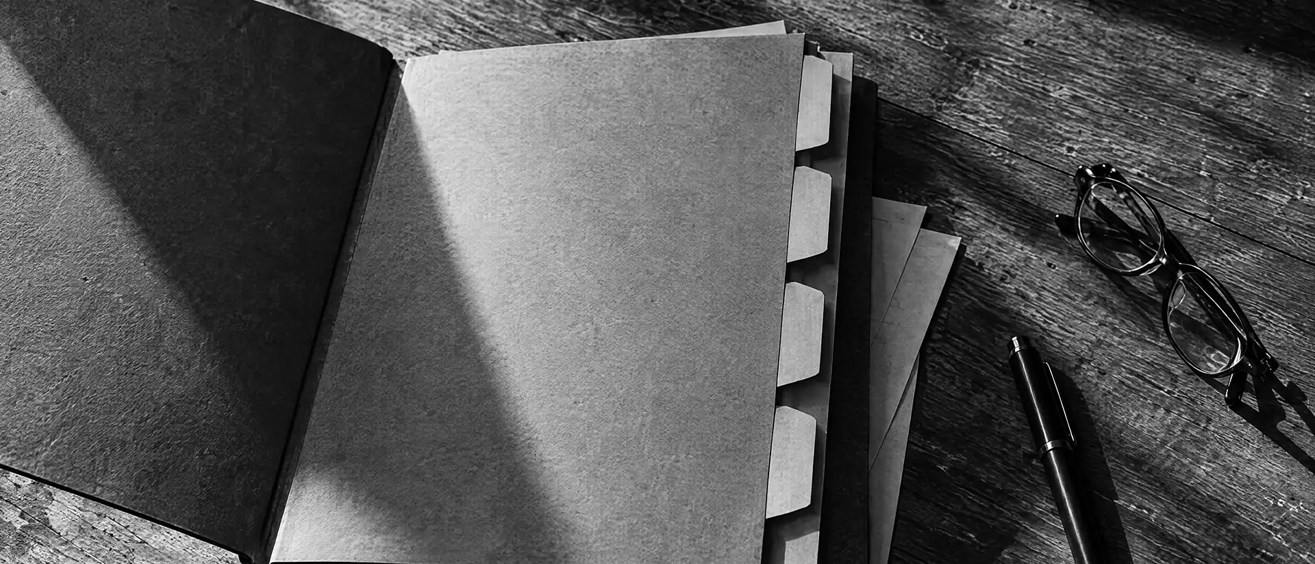 Aufgeschlagene Papiermappe mit losen Blättern und Reitern auf einem Holztisch, daneben Brille und Stift im harten Seitenlicht