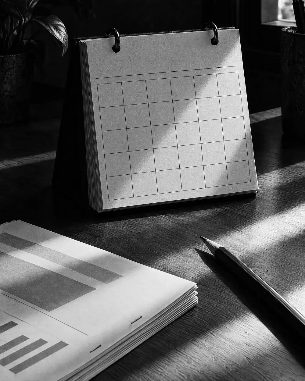 Aufgestellter Tischkalender mit leerem Monatsraster, davor Berichte und ein Bleistift