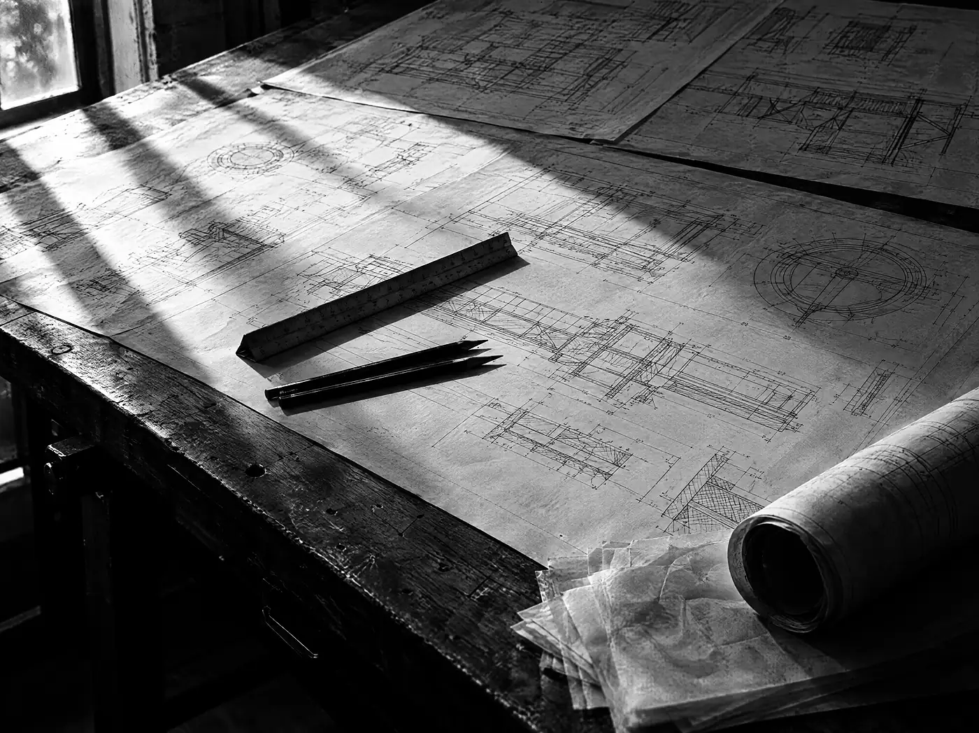 Zeichentisch mit ausgebreiteten technischen Plänen, dreieckigem Maßstabslineal und zwei Bleistiften im harten Seitenlicht