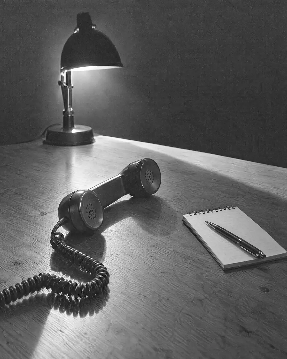 Telefonhörer auf einem Holztisch, daneben Notizblock und Stift, eine eingeschaltete Lampe als einzige Lichtquelle