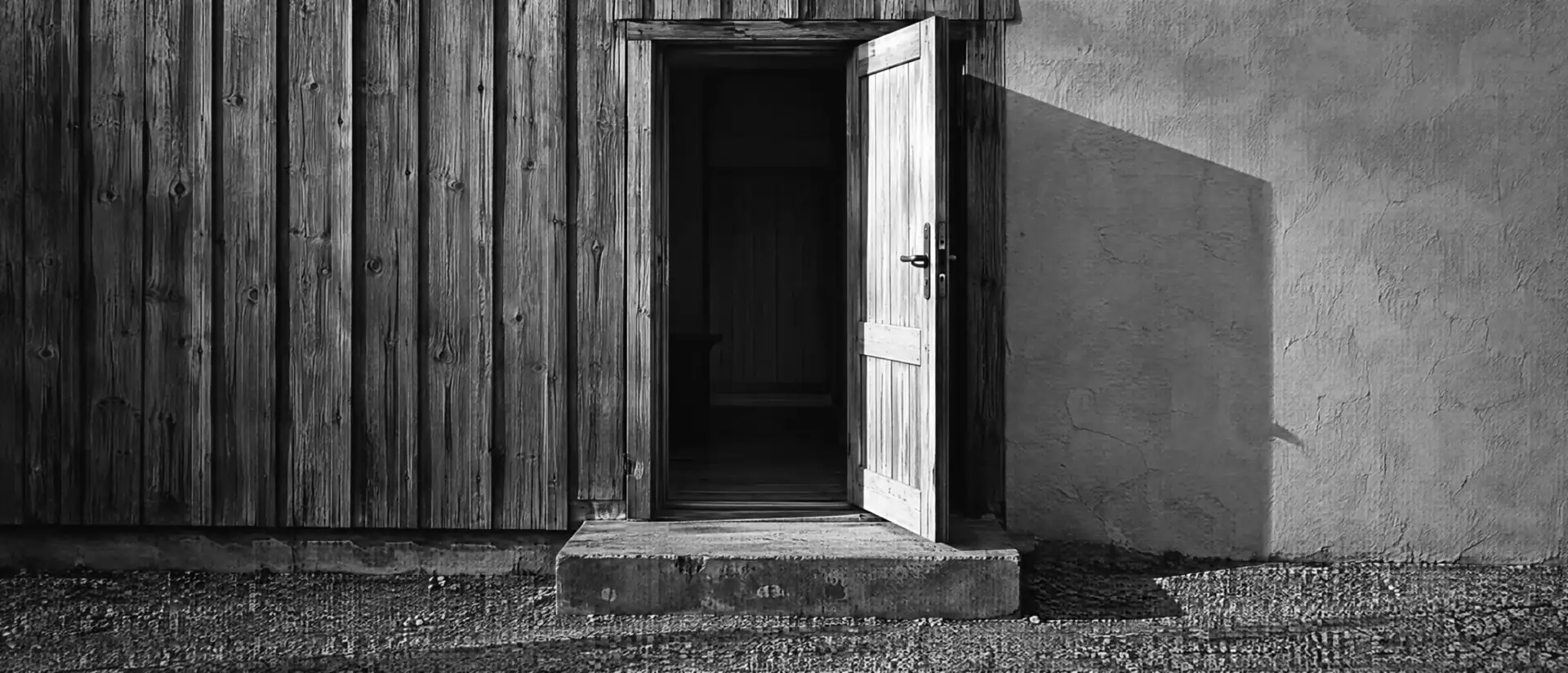 Eingangstür und verwitterte Holzfassade eines kleinen Betriebs, davor eine Stufe und ein Kiesweg im harten Seitenlicht