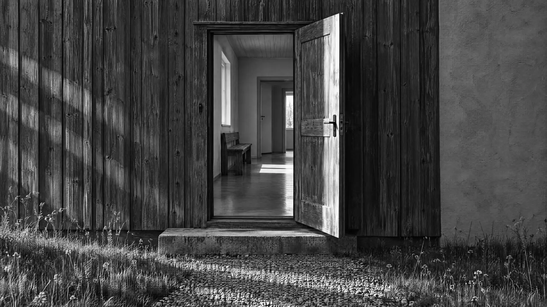 Geöffnete Holztür in der verwitterten Holzfassade eines kleinen Betriebsgebäudes, im hellen Flur steht eine Holzbank, davor Kiesweg mit Wildgras